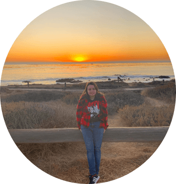 Jessica Heyle wearing a red shirt and black t-shirt standing in front of a wooden gate smiling, the ocean is in the background with a beautiful sunset.