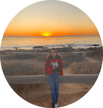 Jessica Heyle wearing a red shirt and black tshirt standing in front of a wooden gate smiling, the ocean is in the background with a beautiful sunset.